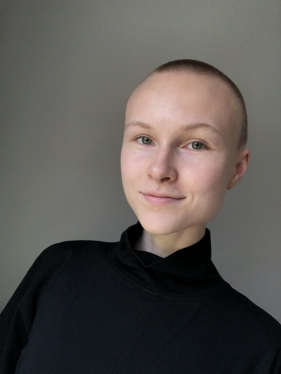 White woman, Hannah Mahr, with a shaved head and a black turtle neck is smiling at the camera.