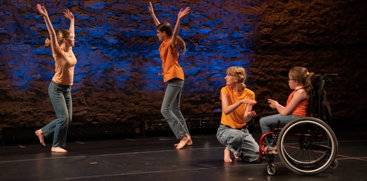Image of two dancers standing with their arms up, and 2 dancers playing patty cake, one in a wheel chair. 
