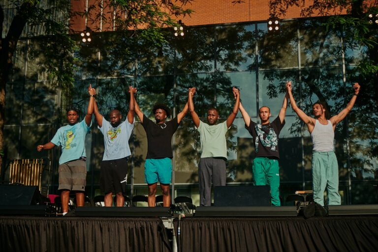Line of black men wearing blue and green colored clothes holding hands with arms held up.