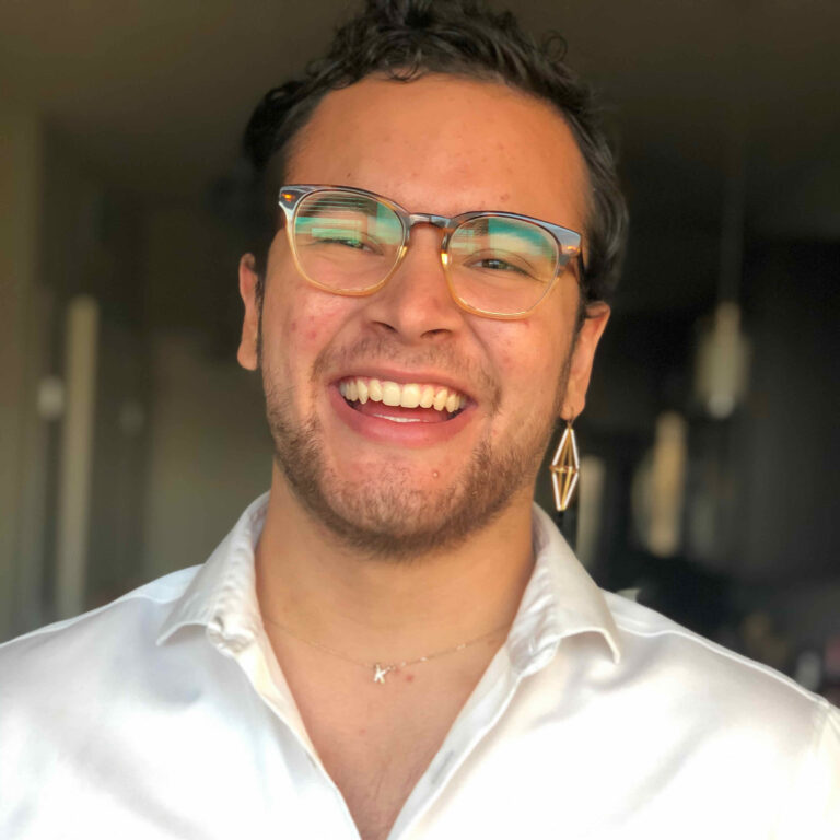 Image of smiling Hispanic man Emio Alvarez, smiling at the camera in a white shirt and wearing one earring.