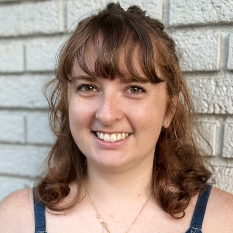 A white woman, Hannah Schwarze, is smiling at the camera in front of a brick wall.