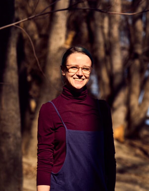 A woman, Meg Singh, standing and smiling in front of trees.